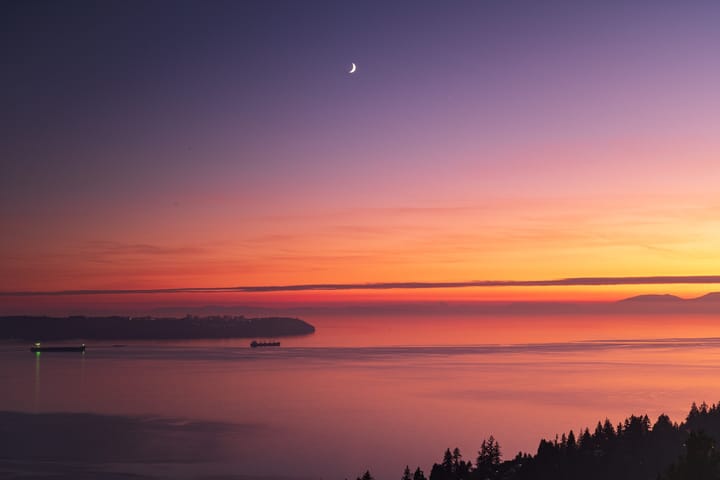 A crescent moon over English Bay at dusk, ships at anchor off Point Grey